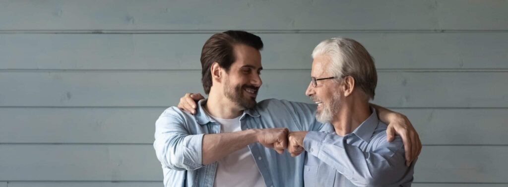 Together we are the power. Bonding mature grandpa young grandson bump fists with smile trust support one another. Happy younger older men relatives friends express unity stand by grey wall.